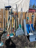 Wide view showing various garden tools arranged upright including rakes, shovels, blue snow shovels, brooms, and rolls of plastic fencing.