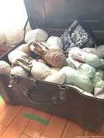 Open military green suitcase box showing assorted yarn skeins in white, beige, and pastel blue shades and a floral bag inside.