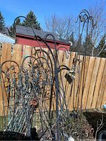Photo showing three tall metal planter hooks with decorative dragonfly accents and curved hooks, along with multiple metal wire trellises and garden decor items in front of a wooden fence outdoors under blue sky.