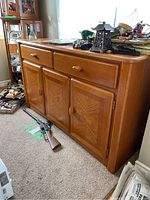 View of entire wooden sideboard showing two upper drawers and three lower doors with inlay wood pattern, placed against a wall on carpeted floor, with miscellaneous items on top and floor around.