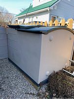 Side and back view of large, beige Rubbermaid resin storage shed with curved dark roof, placed on gravel by a wooden fence.