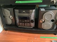 Full view of RCA stereo system with main unit and two speakers on either side, placed on black shelf on wooden floor.