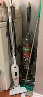 Photo of the Bissell Featherweight stick vacuum, Hoover WindTunnel 3 upright vacuum, and Swiffer mop standing next to a washing machine.