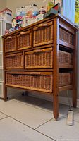Front-left angle showing 6 wicker drawers in a wood frame. Drawers have vinyl handles. Chest placed on tiled floor with items stored on top.