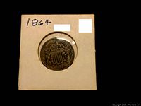 Front side of the 1864 2 Cent Coin showing the shield detail and date, placed in a square cardboard holder with handwritten '1864'