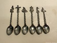 Top view of six souvenir spoons showing detailed figure handles including a seated man, windmill, ship, and others.