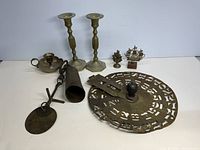 Full lot image showing two brass candlesticks, brass stencil wheel, brass cowbell, chamberstick, pendulum weight, and two decorative pieces.