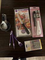 Full overview of the baking bundle laid out on a dark surface. Includes decorating kits in packaging, measuring cups, spatulas, candy thermometer, and silicone brush and tongs.