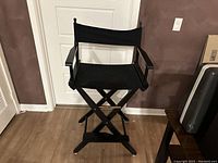 Black wooden director's chair front view, showing X-style legs, armrests, fabric seat and backrest, in a room with wooden floor and white door.