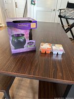Metal ScentSationals fragrance warmer in original purple box, unopened, with two wax melt trays placed alongside on wooden table.