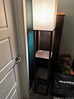 Floor lamp standing upright in corner next to a white door with two open wooden storage shelves, top shelf showing wood grain finish and fabric square lampshade turned on.