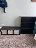 Photo showing small wooden book shelf with three shelves standing upright next to a lower storage shelf with three fabric bins in pastel colors (two blue, one beige)