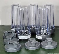 Five clear blending cups arranged upright with separate gray travel lids placed in front, showing the stainless steel blades at the base of each cup.