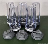 Five clear plastic blender cups each with stainless steel blending blade base, arranged upright in a row. In front of the blender cups, three gray travel lids with flip-top spouts are displayed.