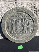 Full front view of the circular antique pewter wall plate showing the detailed embossed scene of a seated royal figure with attendants and a patterned edge.