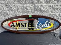 Front view of oval mirror with Amstel Light branding and dark brown frame against brick wall.