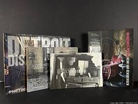 Four photography monograph books arranged standing and stacked: Detroit Disassembled, Abandoned Places, Timeless Islands, Untouched.