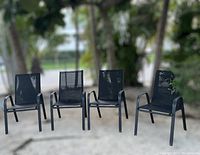 Four black outdoor chairs arranged side by side outdoors on a concrete surface with trees in the background.