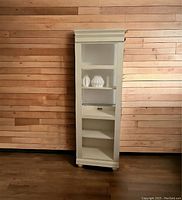 Photograph of narrow solid wood bookcase against wood panel wall. The piece is tall with clean lines and a natural wood finish.