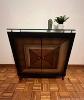 Front view of the dark wood cabinet with two doors and glass top, showing geometric square pattern on doors and chrome metal pegs supporting glass top.