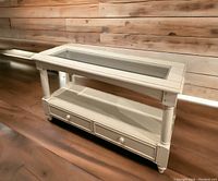 White wood console table with glass inset top, two drawers, and lower shelf, shown on wood floor and in isolated view with black background and measurements overlay.