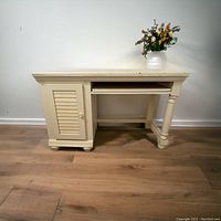Full view of cream-colored solid wood computer desk with storage cabinet and keyboard tray closed, placed against a wall with a floral arrangement on top.