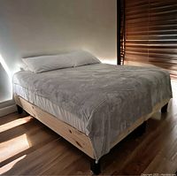 King size bed frame with mattress covered with gray bedding and pillows, placed in a room with wooden flooring and partially closed blinds.