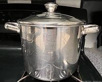 Stainless steel quart pot with glass lid on stovetop, showing reflective surface and dual handles.