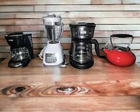 Photo of five kitchen appliances including two coffee makers, one blender, one red tea kettle, and one mixer on wooden surface