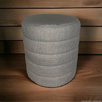 Full view of round ottoman showing textured light gray soft padded fabric and cylindrical shape with horizontal padded sections.