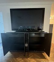 Front view of black wood TV cabinet with flat screen TV on top. Cabinet doors open showing interior shelves.