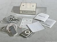 Apple iPod Shuffle with earbuds, charger, plastic case, documentation, and decals shown on flat surface.