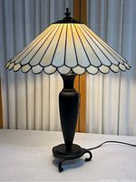 Full view of the lamp showing the black metal base and illuminated scalloped stained glass shade on a white surface with curtain background.