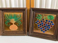 Pair of hammered copper kitchen wall art panels with enameled designs inside dark wood frames, side by side on white surface