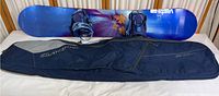 Full view of the Vista Ride snowboard with attached blue bindings laid above the navy blue and grey Dakine carry bag on a surface.