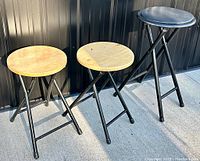 Photo showing all 3 folding stools side by side: two with wood seats and one with black vinyl upholstered seat, all with black metal legs.