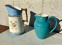 Photo showing two porcelain pitchers on a concrete surface against a wall, one floral decorated with light blue and gold, the other solid teal blue