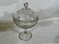 Front angle view of clear antique pressed glass compote with lid, showing intricate geometric patterns on lid and bowl, pedestal base, faceted knob on lid.