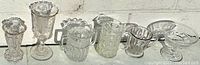 Wide view of seven clear pressed glass pieces including a tall vase, goblet, compote dish, handled creamer and sugar bowl, and other small vessels arranged on a flat surface.
