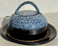 Butter dish dome lid with speckled blue and brown glaze and blue handle on glossy dark brown circular plate.