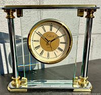 Front view of the Circa floating brass and glass mantle clock showing the clock face and supporting brass columns and base.