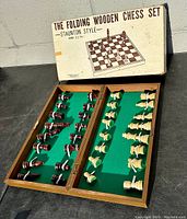 Image showing the folding wooden chess board open with all chess pieces arranged inside on the green felt surface. Box with printed label 'The Folding Wooden Chess Set Staunton Style' in background.