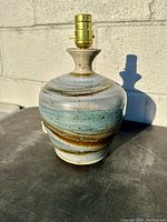Photo of handcrafted stoneware pottery lamp with swirled blue, brown, and beige glaze on a surface against a beige wall, showing the full lamp body and socket.