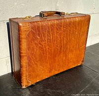Closed view of the brown leather briefcase showing textured, worn leather surface and brass hardware.