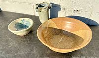 Three signed stoneware pottery pieces: two bowls (brown speckled shallow bowl and small beige bowl with blue-green interior) and one tall cylindrical vase with star-shaped top in grayish-blue glaze.