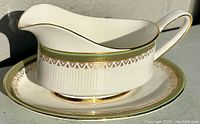 Side-angle photo of bone china gravy boat and matching underplate with green and gold decorative borders, ribbed design on lower half, gold trim on edges and handle.