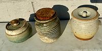 Three lidded pottery jars arranged in sunlight on a concrete surface, showing differing shapes, glazes, and a wooden lid on the middle jar.