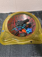 Yellow transparent plastic Beyblade Burst stadium containing Beyblade tops, launchers, and accessories.