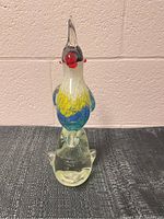 Front view of the blown glass bird figurine showing clear glass base with multicolored body featuring blue, yellow, and red details, placed on dark textured surface against beige wall.