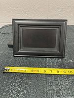Front view of the digital picture frame showing the screen and frame border. A measuring tape in front shows approximately 8 inches width.